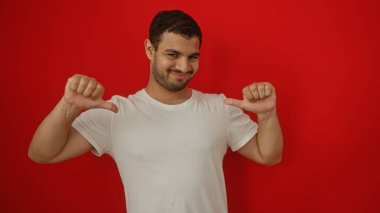 Yakışıklı, İspanyol asıllı, soyutlanmış, kırmızı arka planda gülümseyen beyaz gömlekli ve iki başparmağıyla kendini gösteren bir adam.