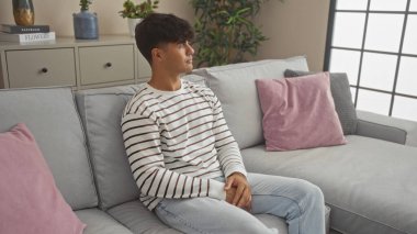 Genç İspanyol bir adam modern bir oturma odasında oturuyor, bir şeyleri yansıtıyor, içinde bitkiler ve minderler olan şık bir iç mekan var..