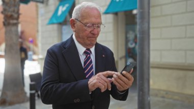 Şehir caddesinde akıllı telefon kullanan, takım elbiseli ve gözlüklü yaşlı beyaz bir adam..
