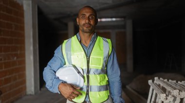 İnşaat alanında güvenlik kaskı tutan yansıtıcı yelek giyen adam endüstriyel ortamda profesyonelliği ve hazır olmayı resmediyor.