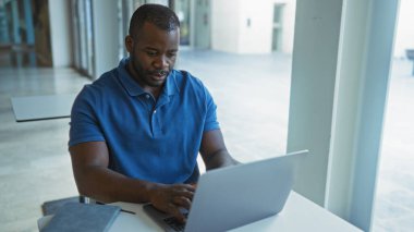 Ofis ortamında dizüstü bilgisayarda çalışan genç bir adam yüzünde odaklanmış bir ifadeyle profesyonel bir çevreyi işaret ediyor..