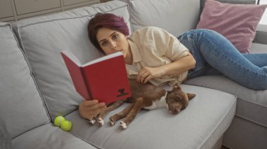 Genç beyaz bir kadın oturma odasındaki kanepede uzanmış köpeğinin yanında kitap okuyor..