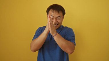 Yakışıklı Çinli adam gülümsüyor ve ellerini çırpıyor sarı bir arka planda izole bir şekilde.