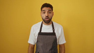 Young hispanic man wears a striped apron over a white shirt, standing isolated against a vibrant yellow background, looking surprised or shocked. clipart