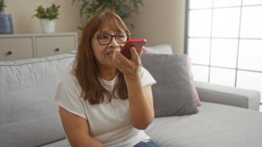 Gözlüklü yaşlı bir İspanyol kadın, bitkiler ve yumuşak tekstil ile çevrili, rahat bir oturma odasında kanepede otururken akıllı telefonuyla konuşuyor..