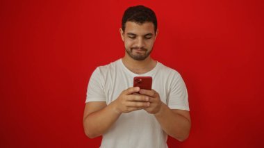 Beyaz tişörtlü genç İspanyol adam izole edilmiş kırmızı arka plan önünde akıllı telefon kullanıyor..