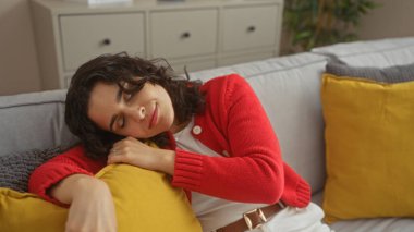 Genç bir İspanyol kadın oturma odasındaki kanepede huzur içinde uyuyor, kırmızı bir kazak giyiyor ve sarı yastıklarla çevrili..