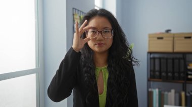 Modern ofis ortamında gözlük takan, uzun siyah saçlı, yeşil bluzlu ve siyah ceketli bir kadın..