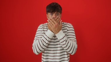 Çizgili tişörtlü, genç, İspanyol bir adam ağzını kırmızı arka planda dikilirken şoka sokuyor..