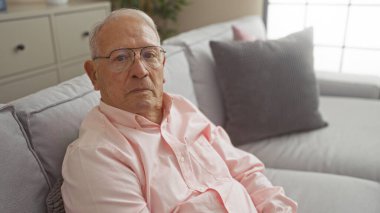Yaşlı, beyaz gözlüklü ve pembe gömlekli bir adam modern bir oturma odasında oturup düşünceli görünüyor..