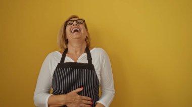 Kısa saçlı, gözlüklü ve parlak sarı arka planda izole edilmiş önlüklü güzel sarışın kadın gülüyor.