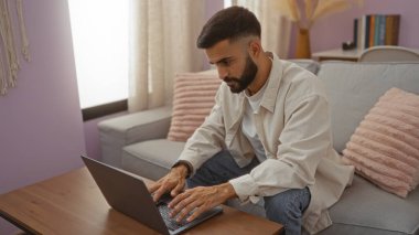 Genç İspanyol adam, pastel dekorlu ve doğal ışıklandırmalı, rahat oturma odasındaki ahşap bir masada dizüstü bilgisayar kullanıyor.