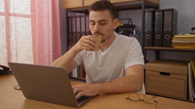 Ofis ortamında sakallı İspanyol bir adam masasında dizüstü bilgisayarla çalışırken kahve içiyor..