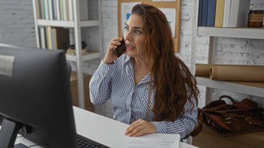 Ofiste telefonla konuşan bir kadın, çalışma masasında kağıtlar ve arka planda raflarda bulunan kitaplarla profesyonel iş ortamını gözler önüne seriyor..