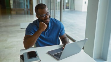 Ofis ortamında dizüstü bilgisayarla çalışan genç bir adam gülümseyen ve gözlük takan, akıllı telefon ve not defteriyle masanın üzerinde.