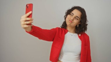 Genç, çekici esmer kadın kırmızı bir telefonla selfie çekiyor, beyaz gömlek ve kırmızı kazak giyiyor, izole edilmiş beyaz bir arka planda..
