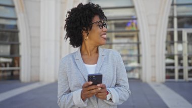 Kıvırcık saçlı ve gözlüklü güzel bir Afro-Amerikalı kadın, şehir caddesinde elinde akıllı bir telefonla gülümsüyor..