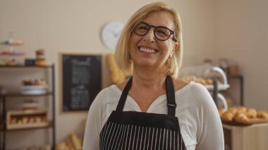 Kısa sarı saçlı, olgun bir kadın fırının içinde gülümsüyor. Arka planda çeşitli hamur işleri ve ekmeklerle.