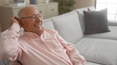 Oturma odasındaki rahat bir kanepede otururken gülümseyen yaşlı beyaz bir adam..