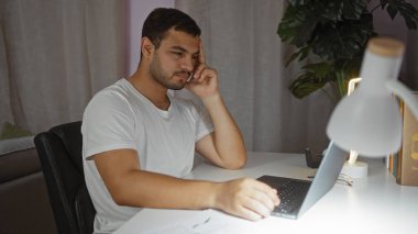 Evde, aydınlık bir oturma odasında dizüstü bilgisayar üzerinde çalışan genç bir İspanyol odak ve konsantrasyon ortamı yaratıyor.