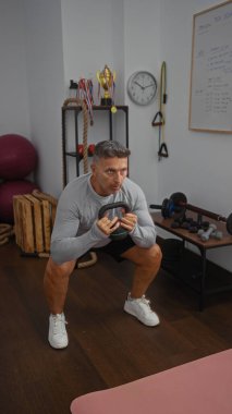 Spor salonunda ağırlıklarla egzersiz yapan, arka planda çeşitli fitness ekipmanları ve motivasyon dekoru olan iyi donanımlı bir jimnastik salonunda kettlebell tutarken çömelen bir adam.