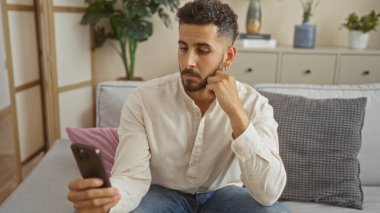 Yakışıklı, Latin asıllı genç bir adam modern oturma odasında oturur, akıllı telefonuna bakar, rahat bir kanepede dinlenir, etrafı bitkilerle ve ev dekoruyla çevrilidir..