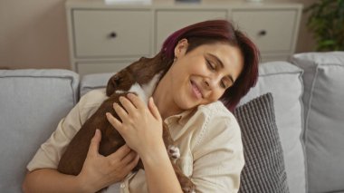 Evindeki rahat oturma odasında rahat bir kanepede otururken köpeğine sevgiyle sarılan güzel beyaz bir kadın..