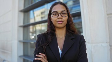 Gözlüklü ve çapraz kollu genç bir kadın şehir ortamında güvenle ayakta duruyor.