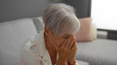 Gri saçlı ve kısa saçlı yaşlı bir kadın bir apartman dairesinde oturuyor. Elleri yüzünü kapatıyor. Stres ve duygusal mücadele hissini yansıtıyor..