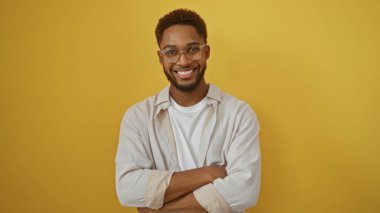 Sarı bir arka plan üzerinde duran kollarını çapraz bağlamış yakışıklı genç adam, rahat kıyafetler ve gözlüklerle güvenle gülümsüyor.