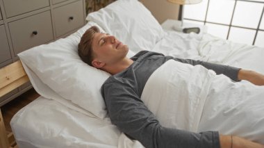 Beyaz çarşaflar ve gri pijamalarla beyaz arka planda, modern bir yatak odasında uyuyan yakışıklı genç adam.