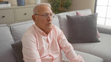 Olgun, gri saçlı, gözlüklü, rahat bir oturma odasında oturan, rahat dekorlu bir adam..