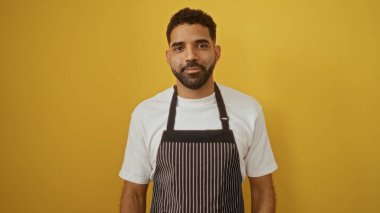 Beyaz gömleğin üzerinde çizgili önlük giyen yakışıklı İspanyol bir adam izole edilmiş sarı bir arka plana karşı duruyor.