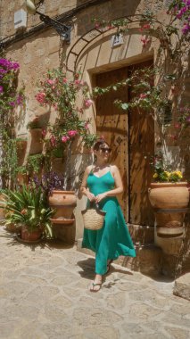 Yeşil elbiseli genç İspanyol kadın Valldemossa, Mallorca, İspanya 'da güneşli bir günün tadını çıkarıyor. Parke sokağında çiçekleri ve bitkileri olan büyüleyici kır evinin önünde duruyor.