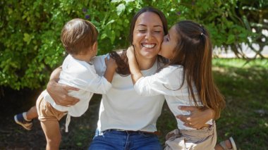 Güneşli park ortamında çocuklara sevgi dolu bir şekilde sarılan, şehir ortamında kızlarıyla oğullarıyla aile bağlarını yakalayan, sıcaklığı ve sevgiyi simgeleyen bir kadın..