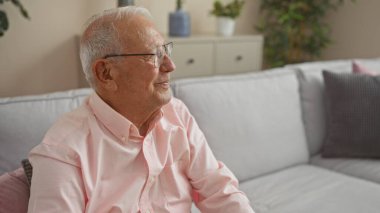 Yaşlı, gri saçlı, beyaz bir adam oturma odasında oturuyor, gözlük takıyor ve pembe bir gömlek giyiyor, halinden memnun ve rahat görünüyor..