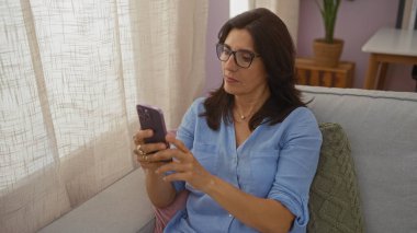Orta yaşlı bir kadın, rahat bir oturma odasında akıllı telefon kullanıyor. Doğal ışık, gözlük ve mavi gömlek takıyor. Arka planda bir yastığın yanında bir kanepede oturuyor..