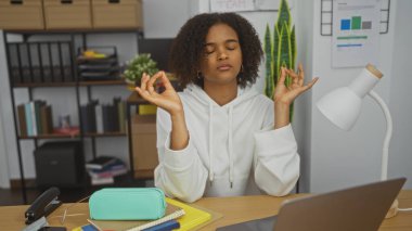 Kıvırcık saçlı, güzel bir Afro-Amerikalı kadın, huzurlu bir ofis ortamında meditasyon yapıyor. Masasında dizüstü bilgisayarı ve kırtasiyesiyle oturuyor. Stres atma ve farkındalık egzersizleri yapıyor..
