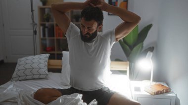 Genç adam rahat bir yatak odasında, hafif ışıklandırma ve içinde kitaplar ve küçük bir lamba bulunan minimalist dekorla geriniyor.