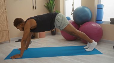 Spor salonunda mavi bir paspasın üzerinde egzersiz yapan genç bir adam. Arka planda spor aletleri ve istikrar topları var.