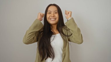 Genç, güzel, Çinli bir kadın beyaz bir arka plana karşı güvenle gülümsüyor ve başarıyı yumruklarıyla kutluyor. Olumlu ve neşe saçıyor..
