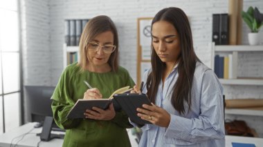 Bir ofiste birlikte çalışan iki kadın, biri not defterine not alırken, diğeri de tablet kullanıyor, profesyonel bir iç mekanda takım çalışması ve işbirliği yapıyor..