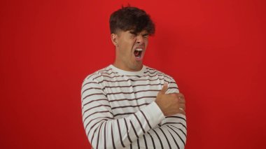 Yakışıklı, genç, İspanyol bir adam kollarını kavuştururken ağzı açık esniyor ve parmaklarını kaldırıyor, parlak kırmızı bir arka plan duvarında izole bir şekilde..