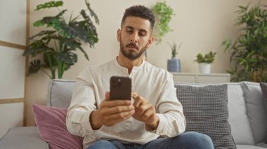 Yakışıklı, genç, İspanyol bir adam rahat bir oturma odasında akıllı telefon kullanarak oturuyor..