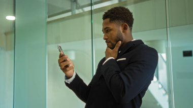 Takım elbiseli genç adam, camdan duvarlar ve parlak ışıklandırmayla çevrili modern bir ofis ortamında dikilirken telefonunu dikkatlice kontrol ediyor..
