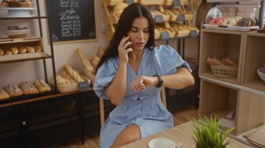 Bir fırında telefonla konuşurken, arka planda hamur işleri ve ekmeklerle saatini kontrol eden genç bir kadın.