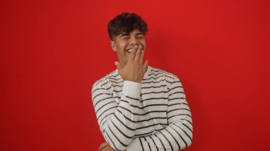 Çizgili tişörtlü yakışıklı İspanyol bir adam gülüyor, parlak kırmızı bir arka plan duvarında izole edilmiş..