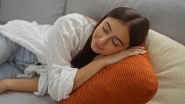 Genç bir kadın, rahat bir oturma odasında turuncu bir yastıkta dinleniyor, beyaz bir gömlek ve kot pantolon giyiyor, esmer saçlı ve huzurlu bir ifadeyle, gündüzleri evde.