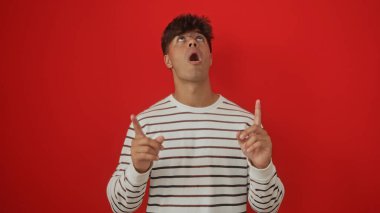 Genç İspanyol bir adam, izole edilmiş kırmızı bir arkaplana işaret ederek sürpriz ya da farkındalık yapıyor..