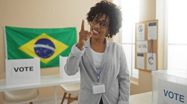 Güzel, kıvırcık saçlı Afro-Amerikan bir kadın işaret parmağını Brezilya bayrakları ve kapalı mekanlarda oy kullanma kabinleriyle neşeyle işaret ediyor..